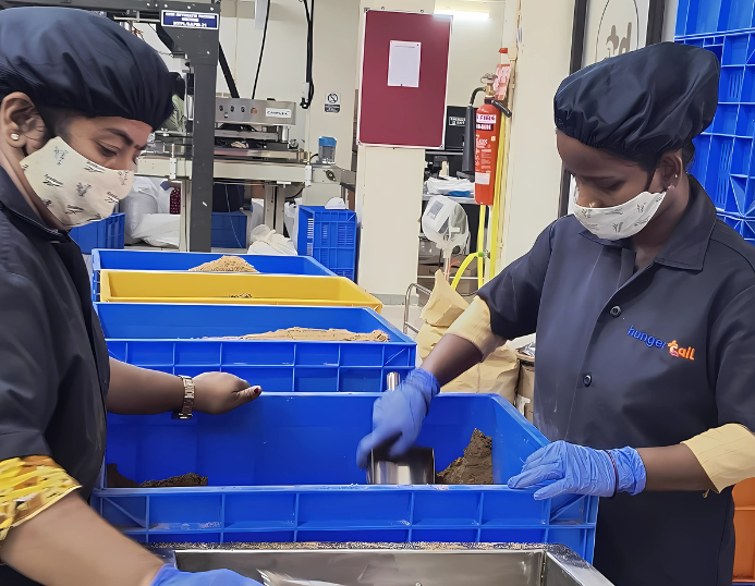 Two women from the Hunger Tail production team wearing masks and gloves while hygienically measuring and packing freshly prepared kibble into blue bins at the factory
